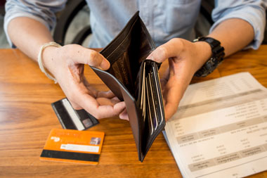 Mann schaut in sein leeres Portemonnaie; neben ihm Rechnung und Kreditkarten auf dem Tisch